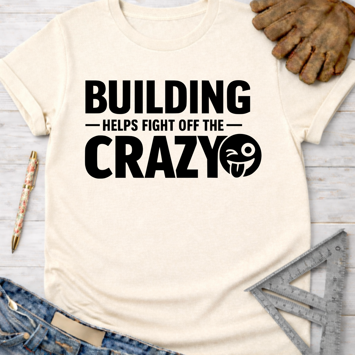 Women's funny woodworking graphic tee shirt for female woodworkers, makers, and DIY builders — Sawdust & Ponytails. Cream short-sleeve cotton tee with "BUILDING HELPS FIGHT OFF THE CRAZY" in black text and a winking emoji.