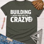 Women's funny woodworking graphic tee shirt for female woodworkers, makers, and DIY builders — Sawdust & Ponytails. Dark gray crew neck t-shirt with "BUILDING HELPS FIGHT OFF THE CRAZY" text and winking emoji.