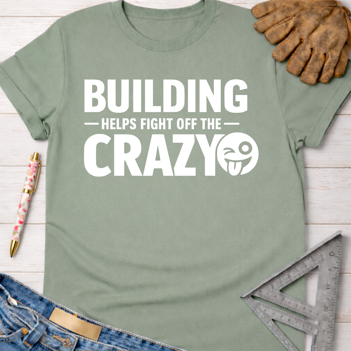Women's funny woodworking graphic tee shirt for female woodworkers, makers, and DIY builders — Sawdust & Ponytails. Sage green cotton t-shirt with "BUILDING HELPS FIGHT OFF THE CRAZY" in white text and winking emoji.