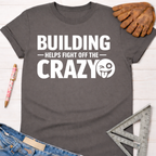 Women's funny woodworking graphic tee shirt for female woodworkers, makers, and DIY builders — Sawdust & Ponytails. Dark gray cotton tee with "BUILDING HELPS FIGHT OFF THE CRAZY" text and winking emoji.