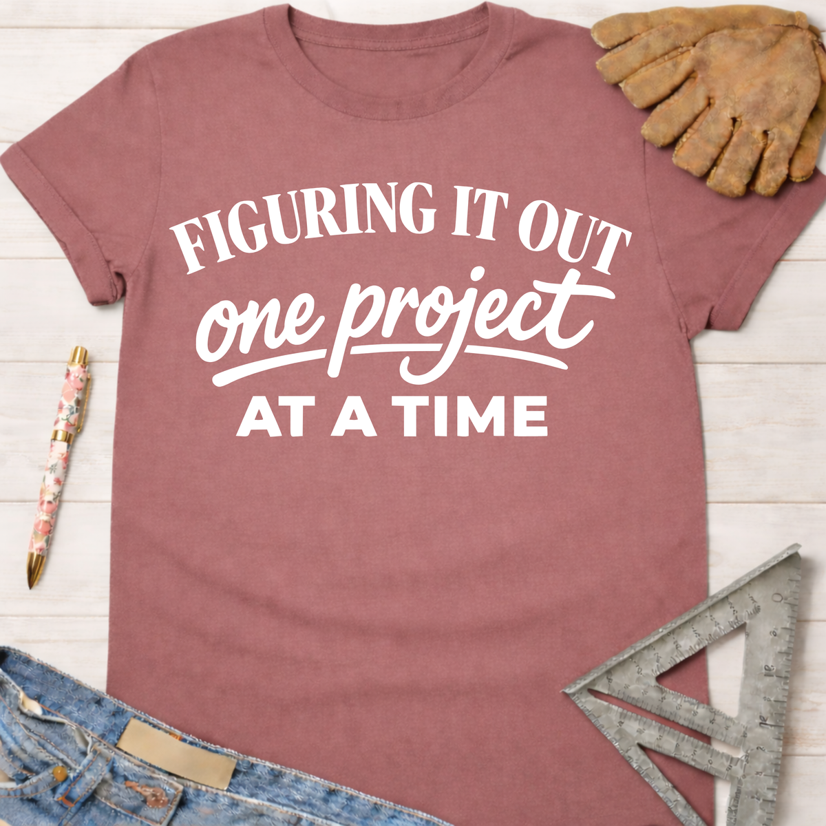 Women's funny woodworking graphic tee shirt for female woodworkers, makers, and DIY builders — Sawdust & Ponytails. Mauve cotton tee with "Figuring It Out One Project At A Time" in white text.