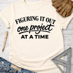 Women's funny woodworking graphic tee shirt for female woodworkers, makers, and DIY builders — Sawdust & Ponytails. Cream t-shirt with "Figuring It Out One Project At A Time" in black text.