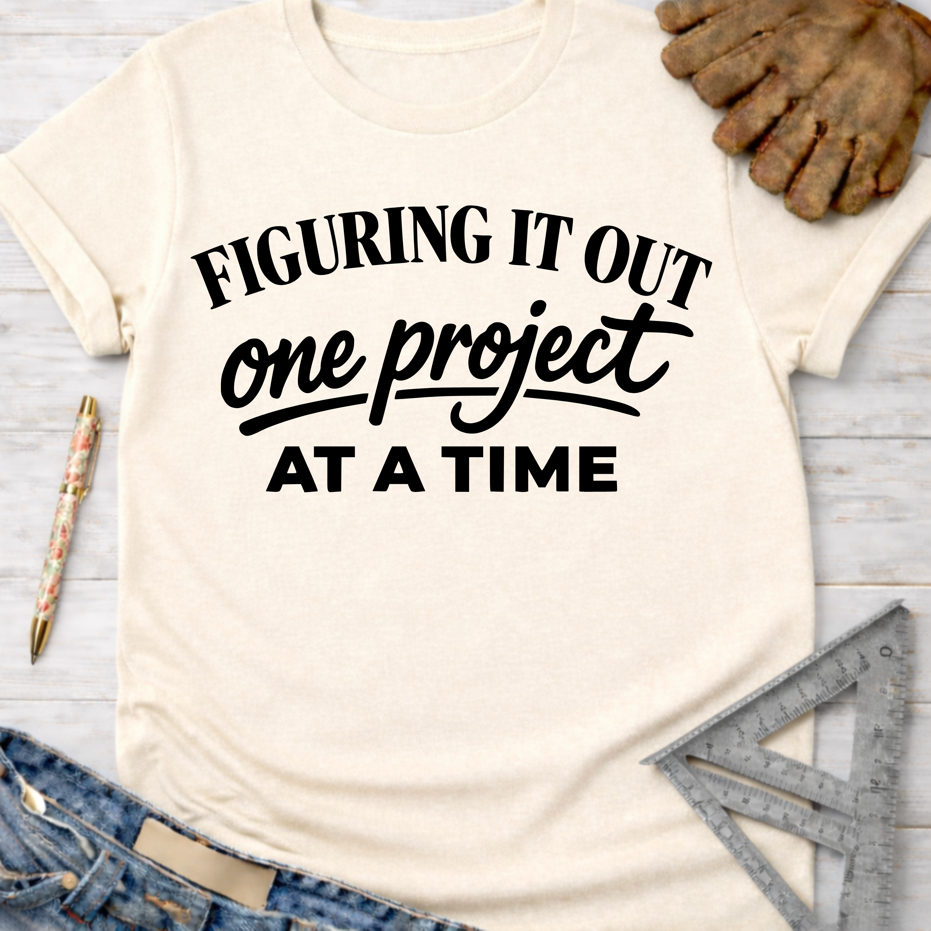 Women's funny woodworking graphic tee shirt for female woodworkers, makers, and DIY builders — Sawdust & Ponytails. Cream t-shirt with "Figuring It Out One Project At A Time" in black text.