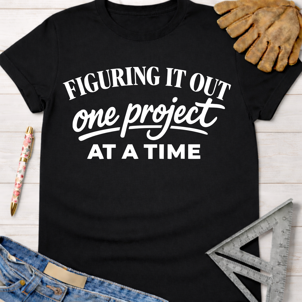 Women's funny woodworking graphic tee shirt for female woodworkers, makers, and DIY builders — Sawdust & Ponytails. Black cotton tee with "Figuring It Out One Project At A Time" in white text.