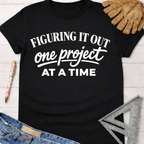 Women's funny woodworking graphic tee shirt for female woodworkers, makers, and DIY builders — Sawdust & Ponytails. Black cotton tee with "Figuring It Out One Project At A Time" in white text.