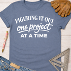 Women's funny woodworking graphic tee shirt for female woodworkers, makers, and DIY builders — Sawdust & Ponytails. Blue cotton tee with "Figuring It Out One Project At A Time" in white text.