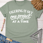 Sage green cotton tee with "FIGURING IT OUT one project AT A TIME" graphic. Women's funny woodworking graphic tee shirt for female woodworkers, makers, and DIY builders — Sawdust & Ponytails.