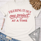 Women's funny woodworking graphic tee shirt for female woodworkers, makers, and DIY builders — Sawdust & Ponytails. Cream short-sleeve t-shirt with "Figuring It Out One Project At A Time" in pink text.