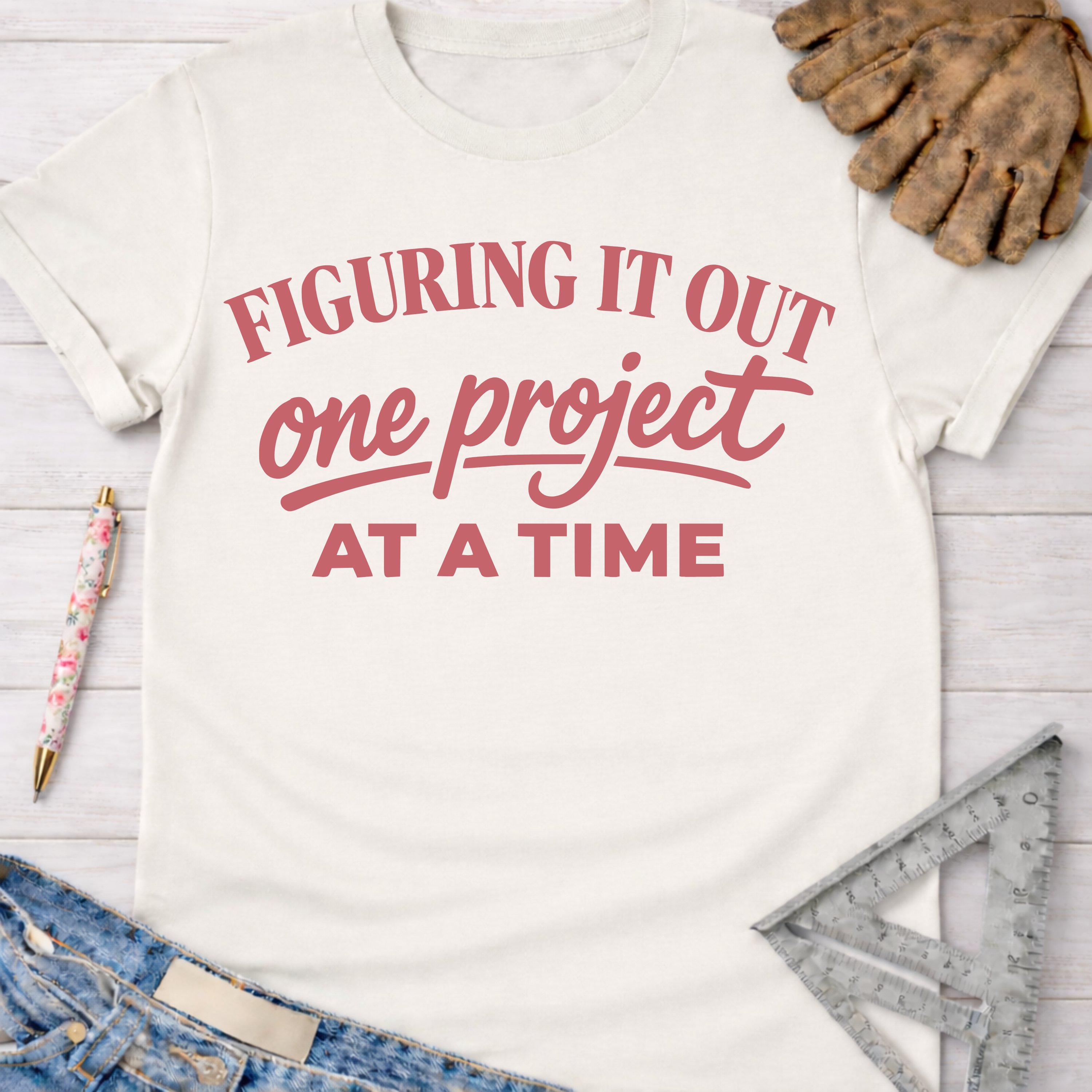 Women's funny woodworking graphic tee shirt for female woodworkers, makers, and DIY builders — Sawdust & Ponytails. Cream short-sleeve t-shirt with "Figuring It Out One Project At A Time" in pink text.