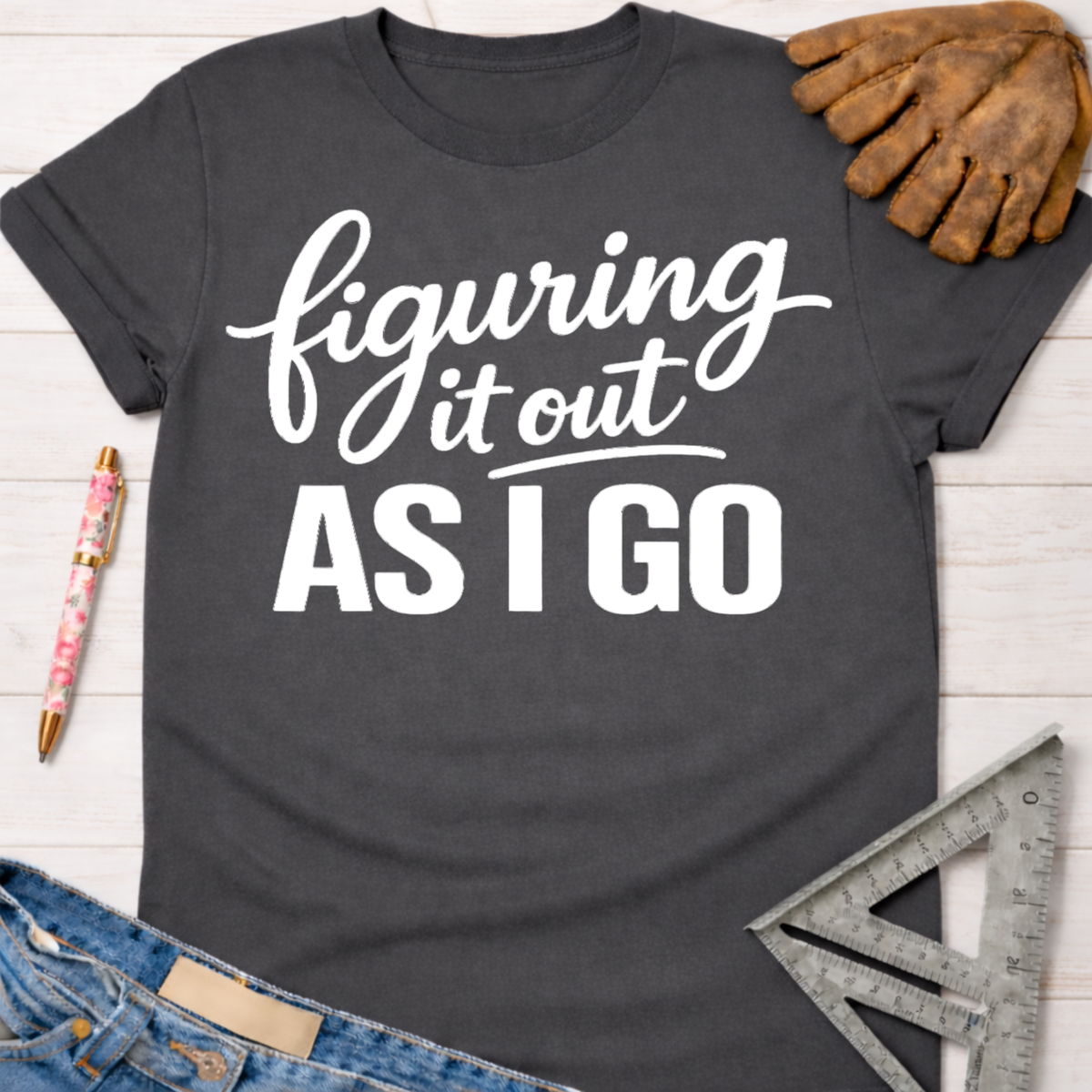 Women's funny woodworking graphic tee shirt for female woodworkers, makers, and DIY builders — Sawdust & Ponytails. Dark gray cotton tee with "figuring it out AS I GO" in white script.