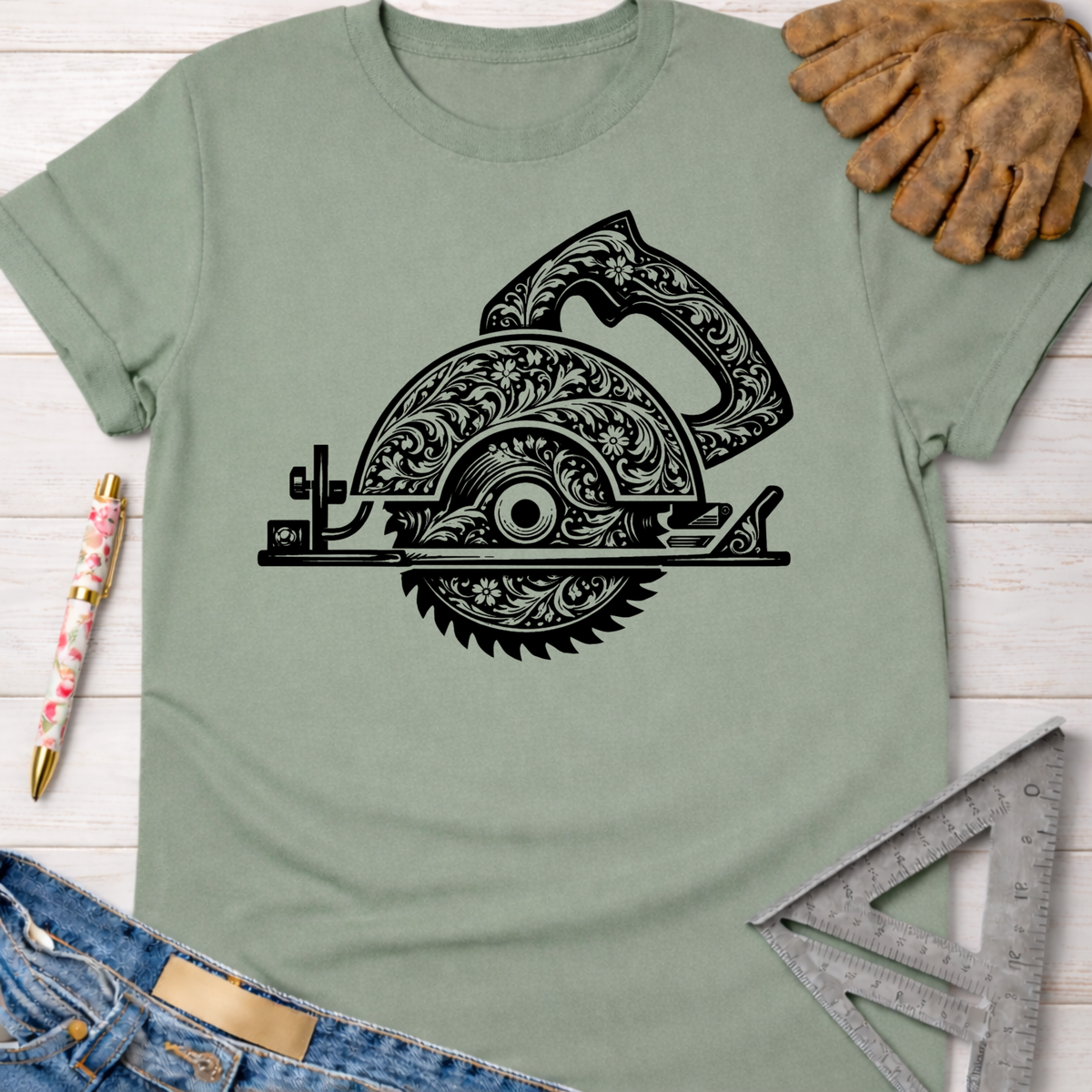 Women's funny woodworking graphic tee shirt for female woodworkers, makers, and DIY builders — Sawdust & Ponytails. Sage green t-shirt with a black floral engraved circular saw graphic, next to jeans, work gloves, and a ruler.