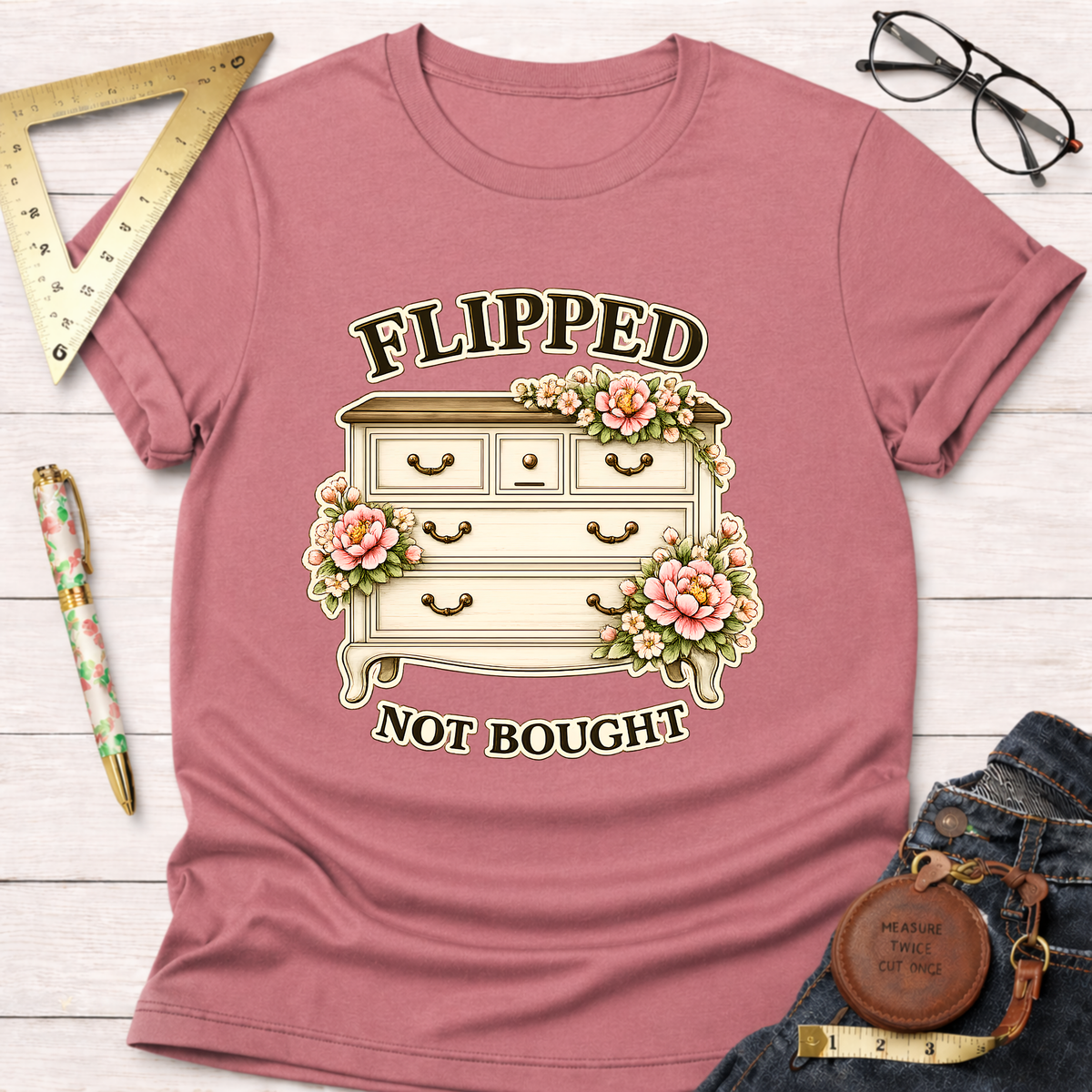 Pink "Flipped Not Bought" t-shirt with a vintage dresser and floral design, styled on a white wooden surface.