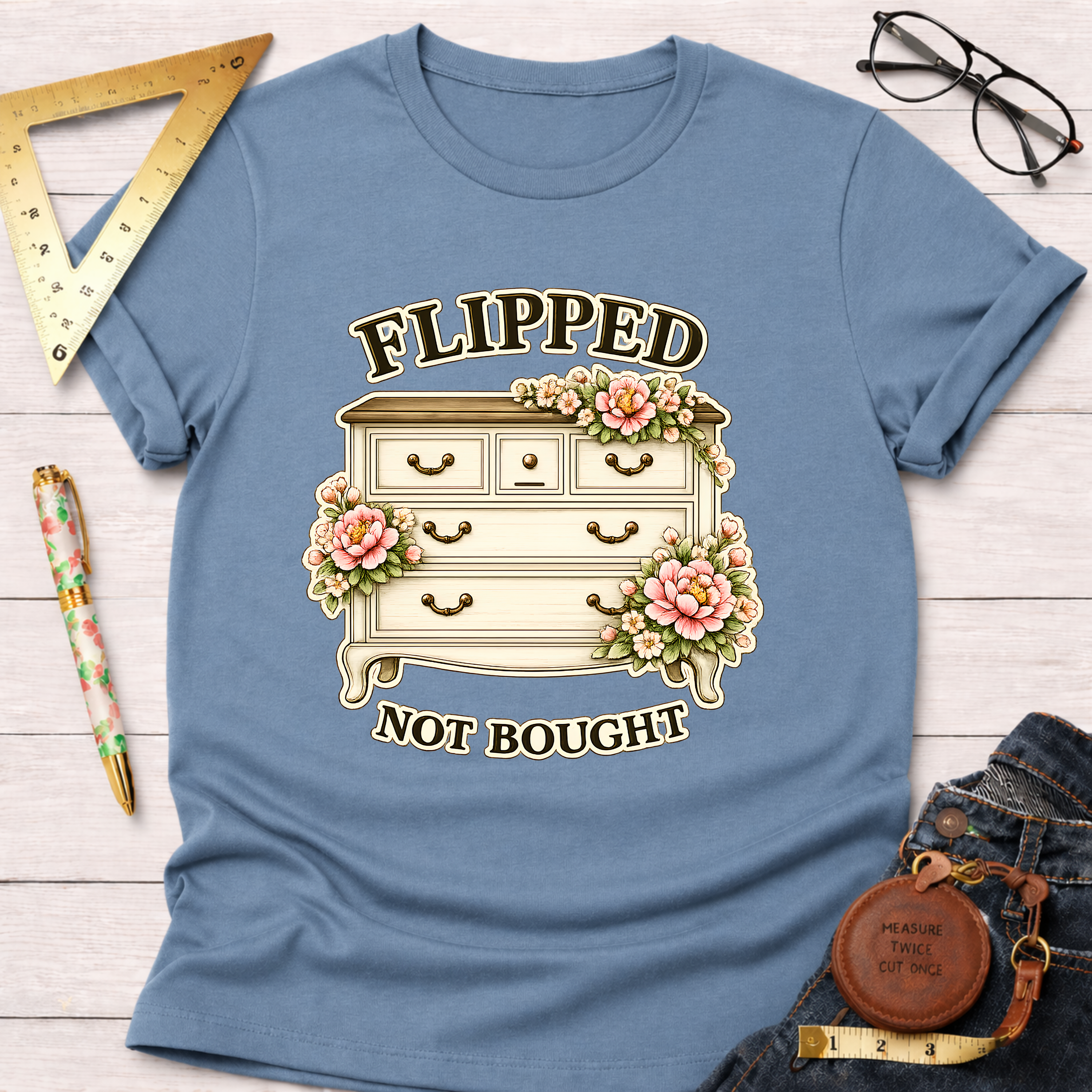 Blue cotton t-shirt with "Flipped Not Bought" text and a vintage dresser adorned with pink flowers.