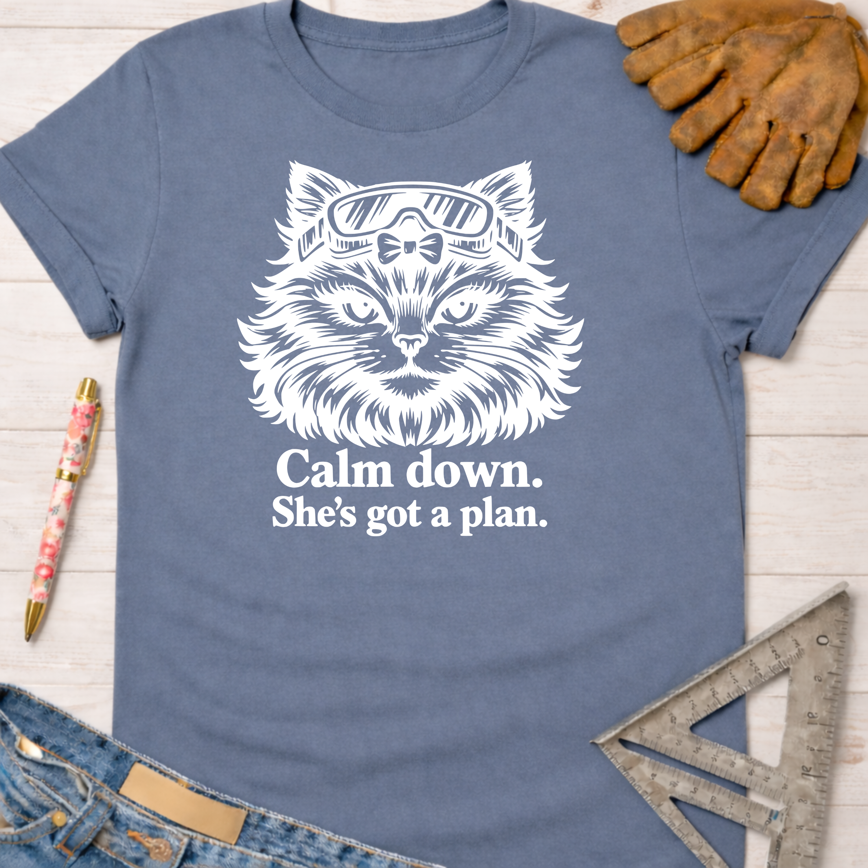 Women's funny woodworking graphic tee shirt for female woodworkers, makers, and DIY builders — Sawdust & Ponytails. Blue cotton tee with white cat in goggles graphic and "Calm down. She's got a plan." text.
