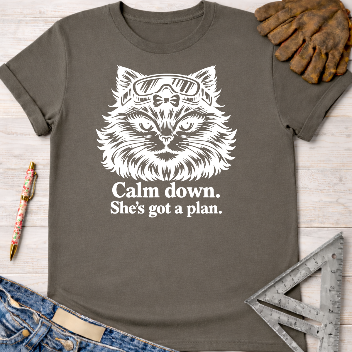 Women's funny woodworking graphic tee shirt for female woodworkers, makers, and DIY builders — Sawdust & Ponytails. Dark gray cotton tee with white cat wearing goggles and text "Calm down. She's got a plan.