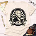 Cream t-shirt with a black graphic of George Washington, "America EST. 1776," and American flags.