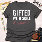 Charcoal gray "Gifted With Skill & Sawdust" t-shirt with white and pink text, folded on a light wood surface.