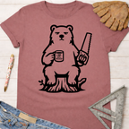 Women's funny woodworking graphic tee shirt for female woodworkers, makers, and DIY builders — Sawdust & Ponytails. Mauve t-shirt with a black graphic of a grumpy bear holding a saw and "I Love Wood" mug, on a white wood background with jeans, a pen, and a square ruler.