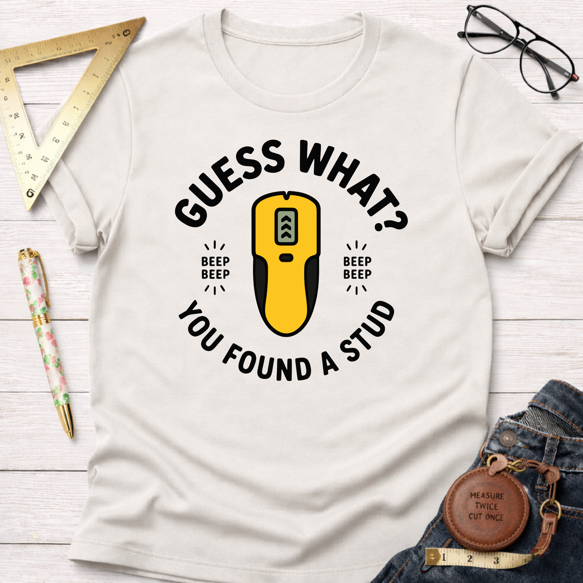 White "Guess What? You Found A Stud" t-shirt with a yellow stud finder graphic, surrounded by carpentry tools.