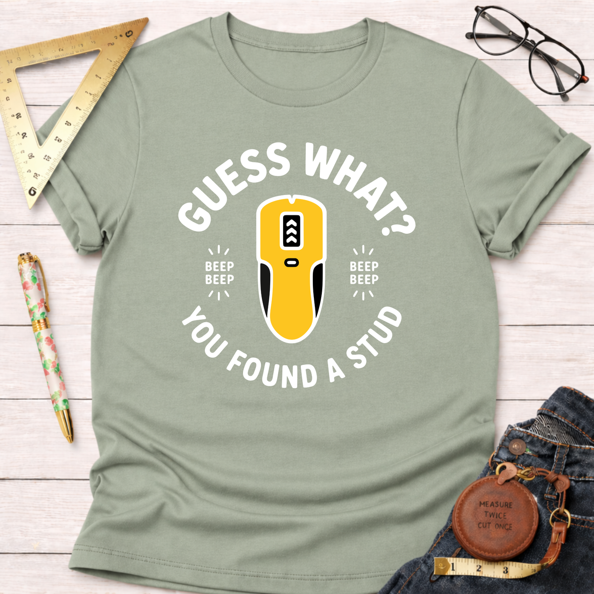 Sage green "Guess What? You Found A Stud" t-shirt with a yellow stud finder graphic, styled with jeans and tools.
