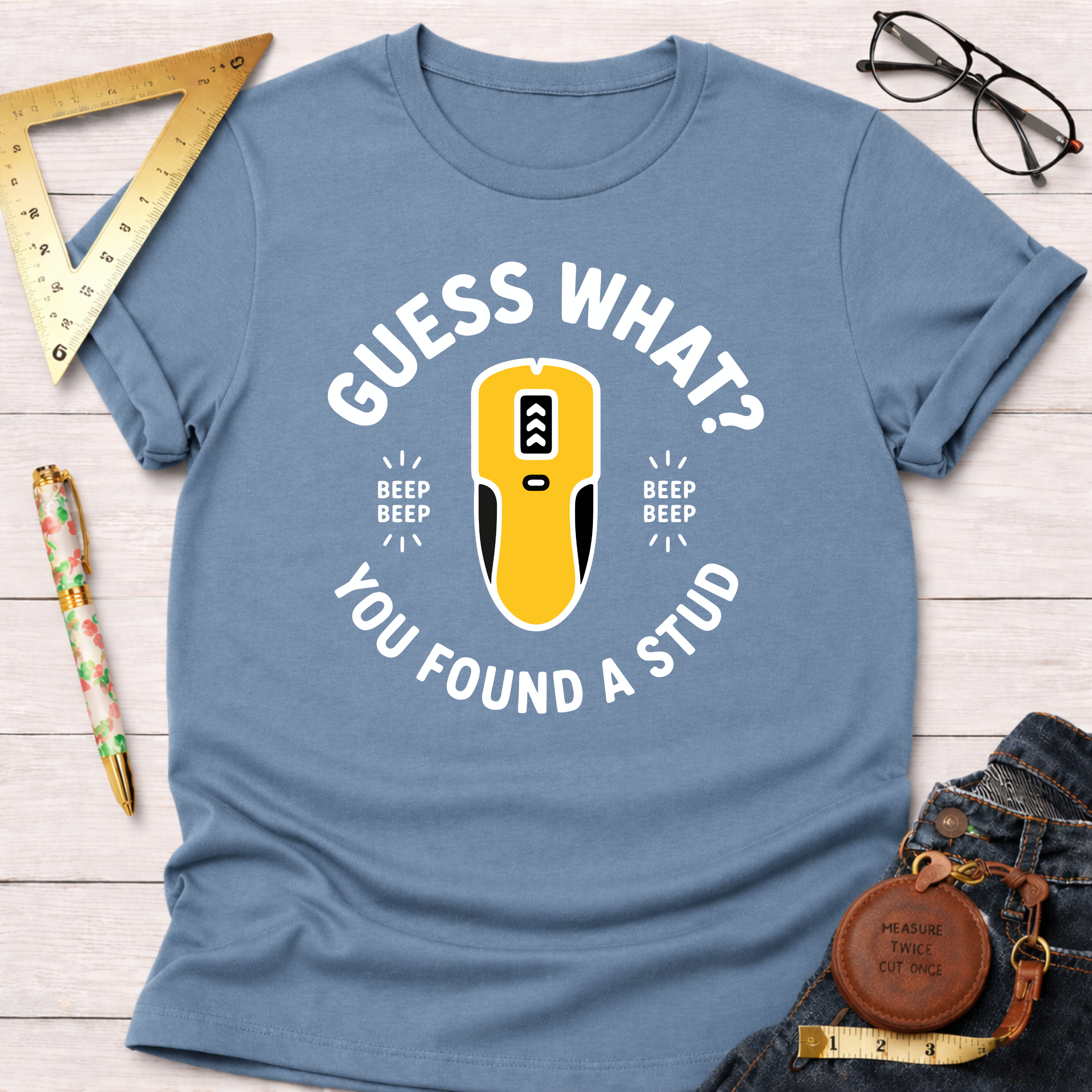 Light blue "Guess What? You Found A Stud" t-shirt with a yellow stud finder graphic, styled with jeans and tools.