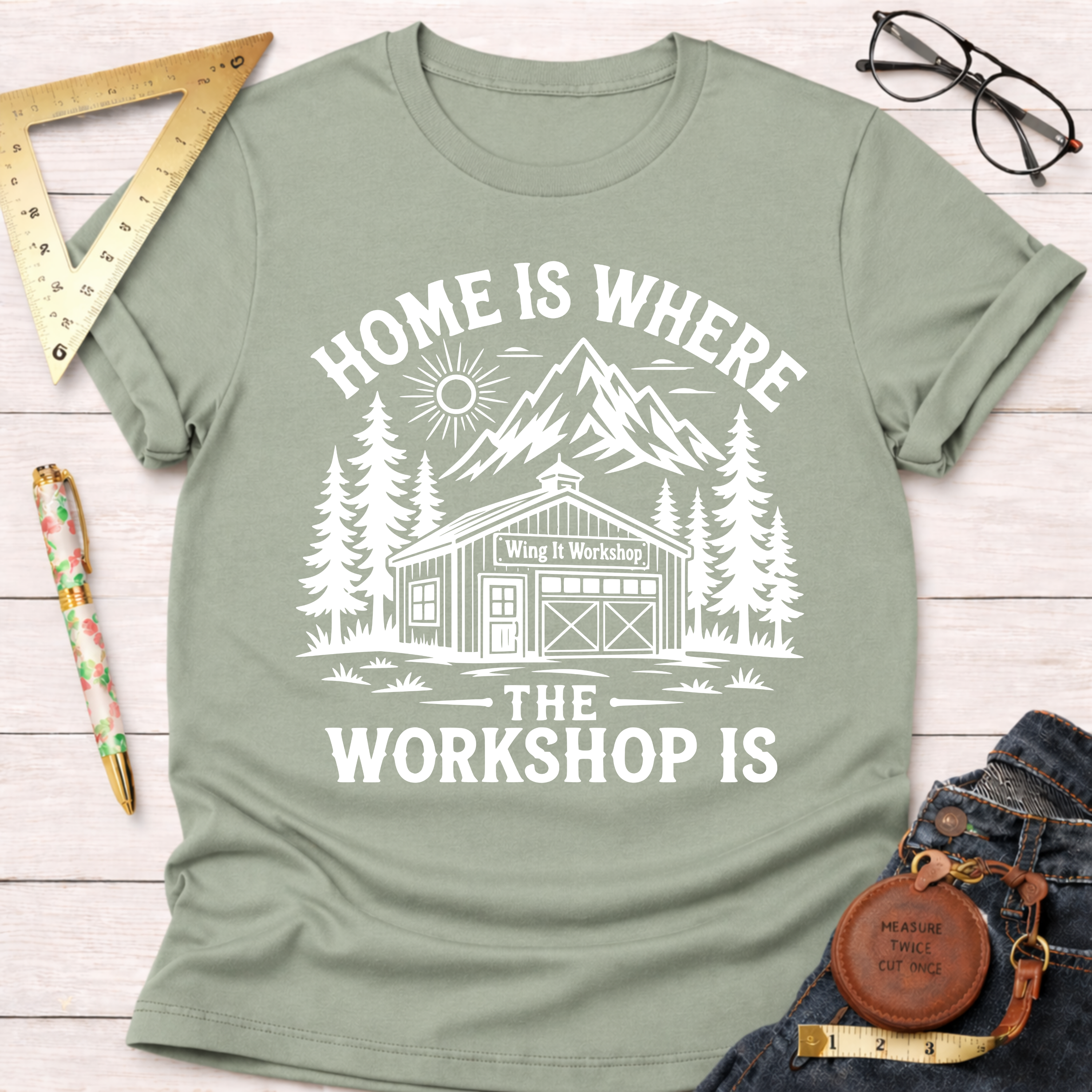 Women's funny woodworking graphic tee shirt for female woodworkers, makers, and DIY builders — Sawdust & Ponytails. Sage green cotton tee with "Home Is Where The Workshop Is" graphic.