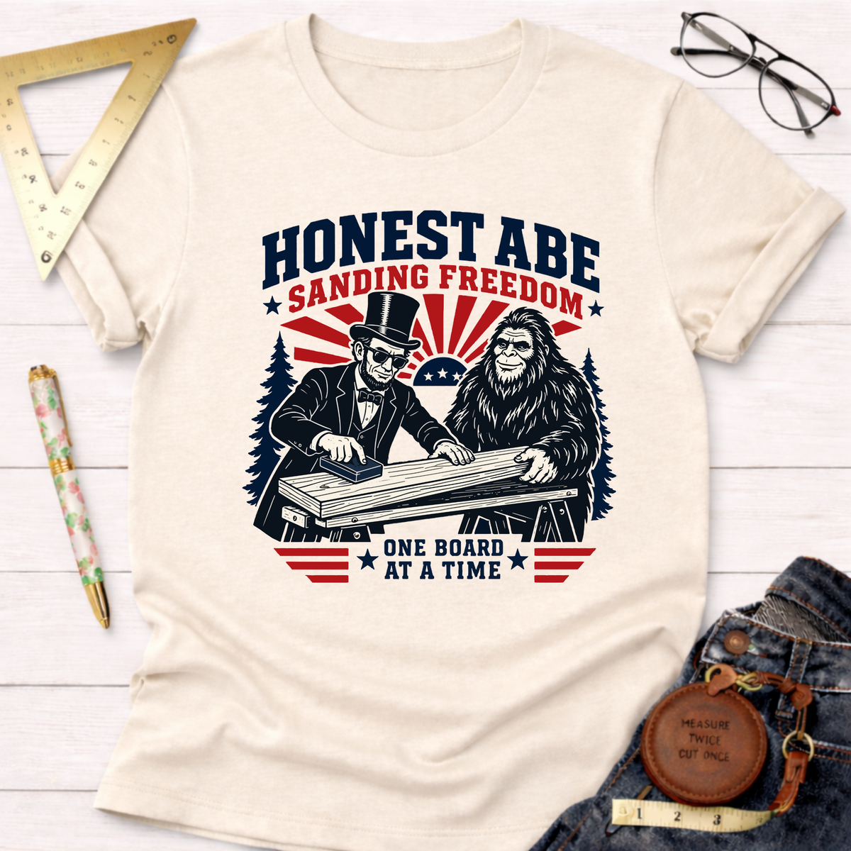 Cream cotton tee with "Honest Abe Sanding Freedom" graphic featuring Lincoln and Bigfoot woodworking.