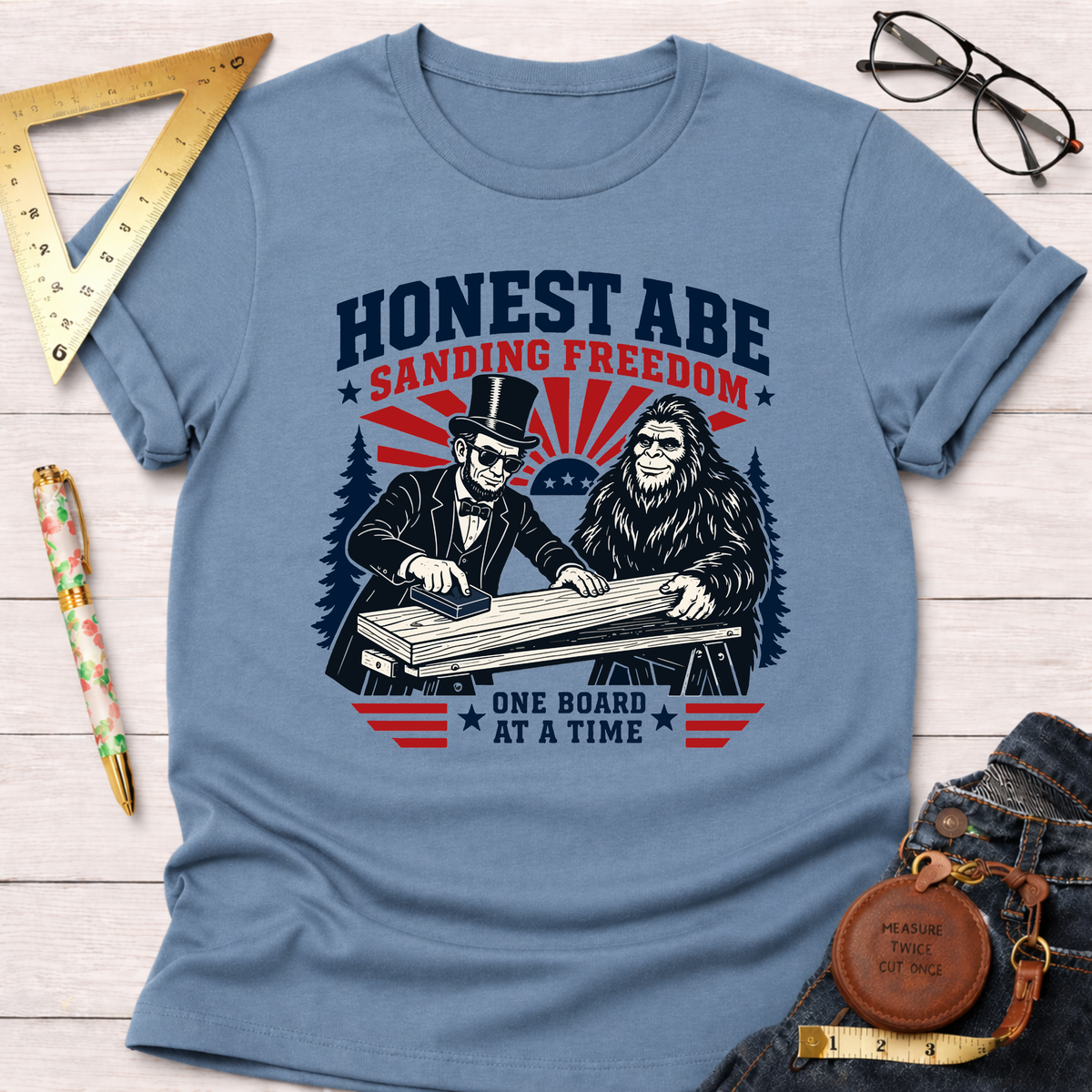 Light blue t-shirt with a graphic of Abraham Lincoln and Bigfoot sanding wood, "Honest Abe Sanding Freedom" text.