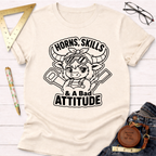 Cream t-shirt with a black graphic of a cute horned cow wearing goggles and overalls, holding tools, with "Horns, Skills, & A Bad Attitude" text.