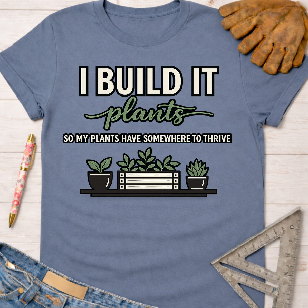 Women's funny woodworking graphic tee shirt for female woodworkers, makers, and DIY builders — Sawdust & Ponytails. Blue cotton tee with "I BUILD IT plants SO MY PLANTS HAVE SOMEWHERE TO THRIVE" graphic and potted plants.