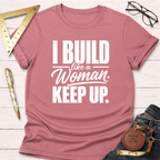 Pink short-sleeve t-shirt with "I BUILD like a Woman KEEP UP." in white text, styled on a white wood surface.