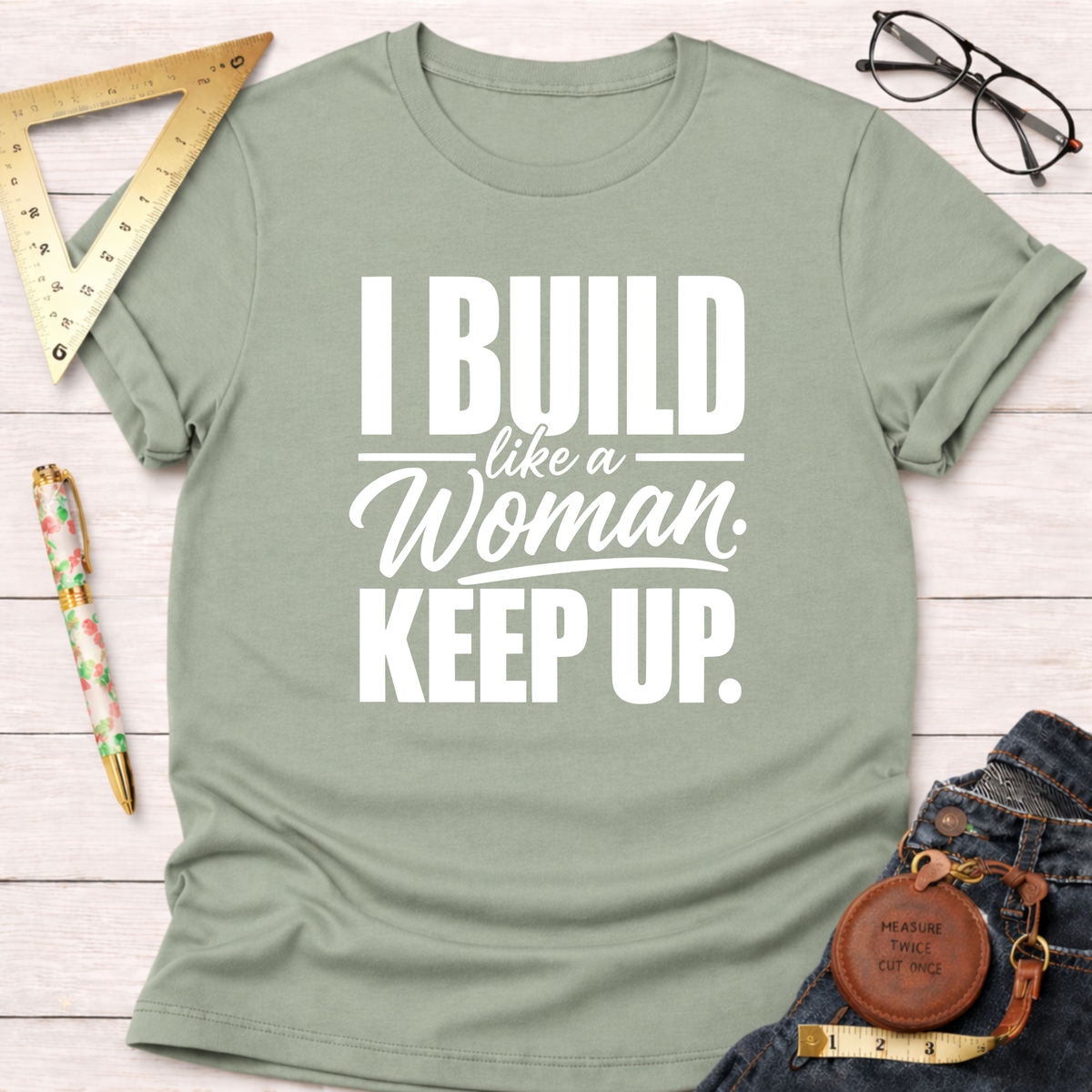Sage green cotton t-shirt with "I Build Like A Woman. Keep Up." in white text, styled on a white wood surface.