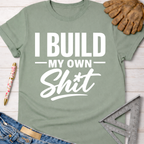 Women's funny woodworking graphic tee shirt for female woodworkers, makers, and DIY builders — Sawdust & Ponytails. Sage green cotton t-shirt with "I Build My Own Shit" in white text.