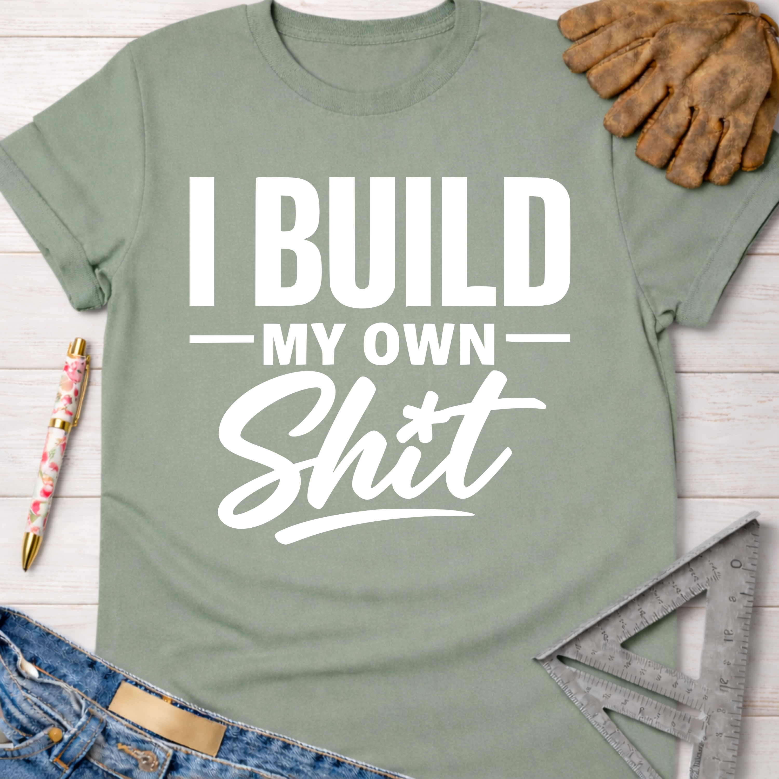 Women's funny woodworking graphic tee shirt for female woodworkers, makers, and DIY builders — Sawdust & Ponytails. Sage green cotton t-shirt with "I Build My Own Shit" in white text.