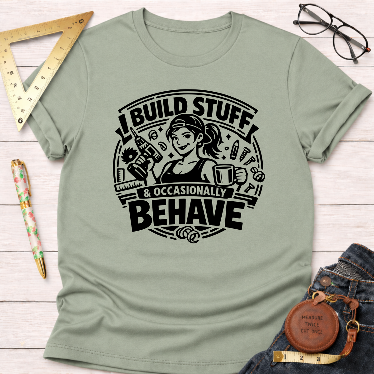 Light olive green cotton t-shirt with "I Build Stuff & Occasionally Behave" graphic featuring a woman with a drill and coffee.