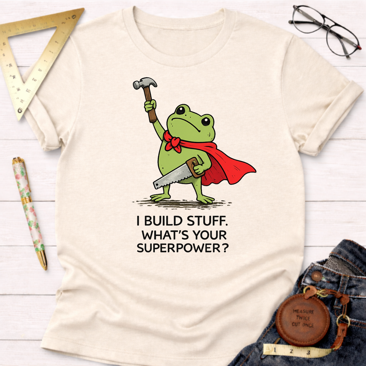 Women's funny woodworking graphic tee shirt for female woodworkers, makers, and DIY builders — Sawdust & Ponytails. Cream cotton tee with a superhero frog holding a hammer and saw, text "I BUILD STUFF. WHAT'S YOUR SUPERPOWER?".