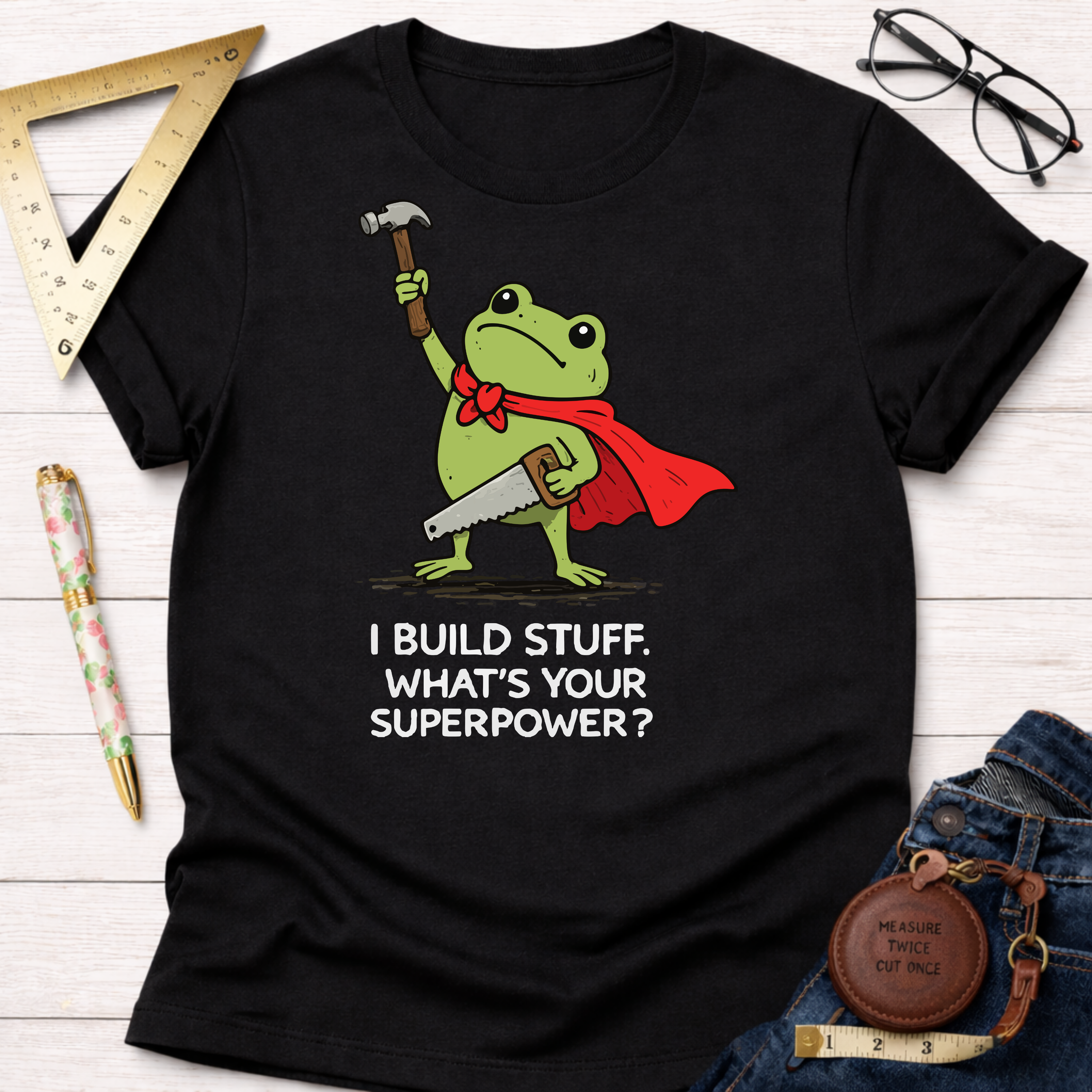 Black cotton tee with a frog in a red cape holding a hammer and saw, "I Build Stuff. What's Your Superpower?" text. Women's funny woodworking graphic tee shirt for female woodworkers, makers, and DIY builders — Sawdust & Ponytails