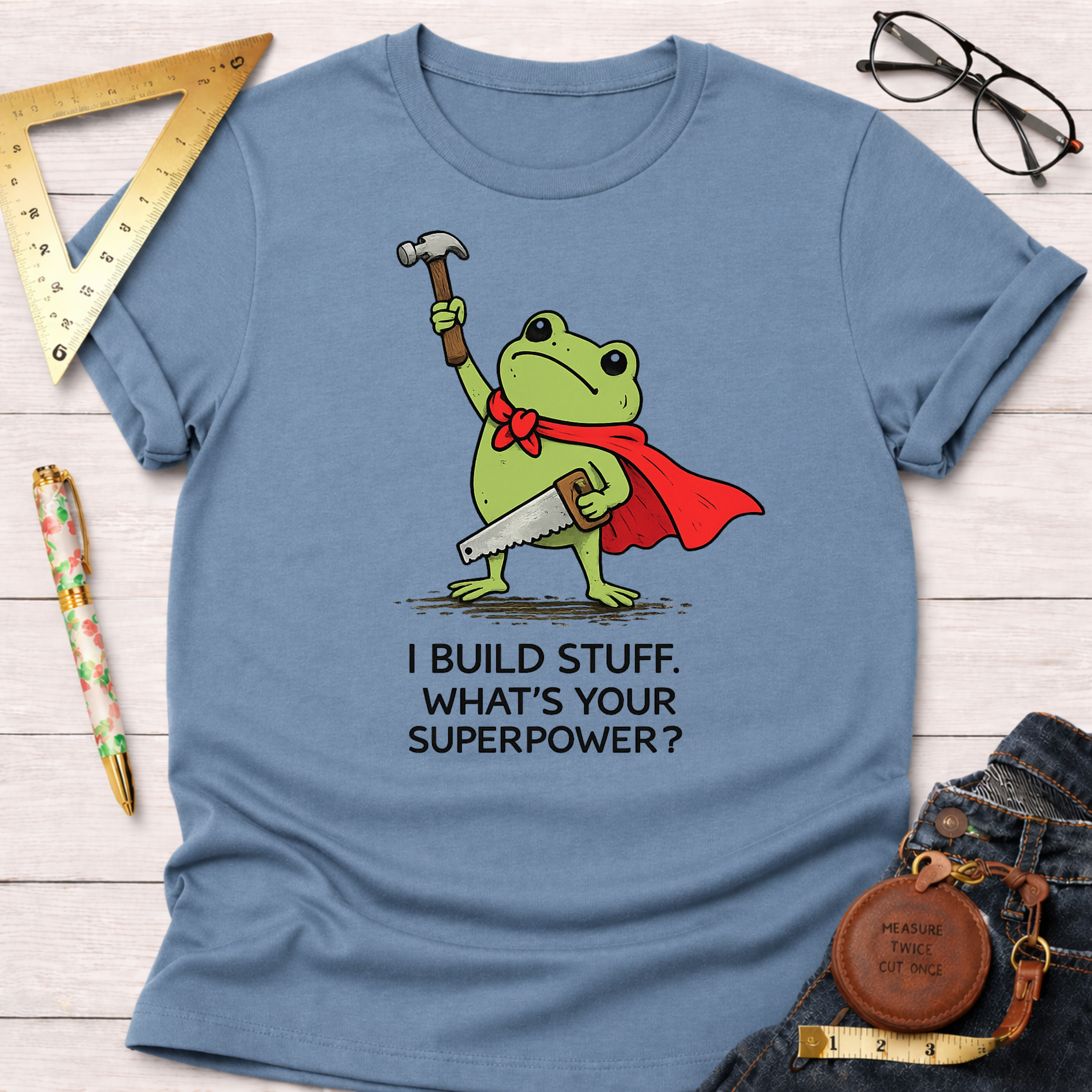 Light blue cotton tee with a frog superhero holding a hammer and saw, "I BUILD STUFF. WHAT'S YOUR SUPERPOWER?" text. Women's funny woodworking graphic tee shirt for female woodworkers, makers, and DIY builders — Sawdust & Ponytails.