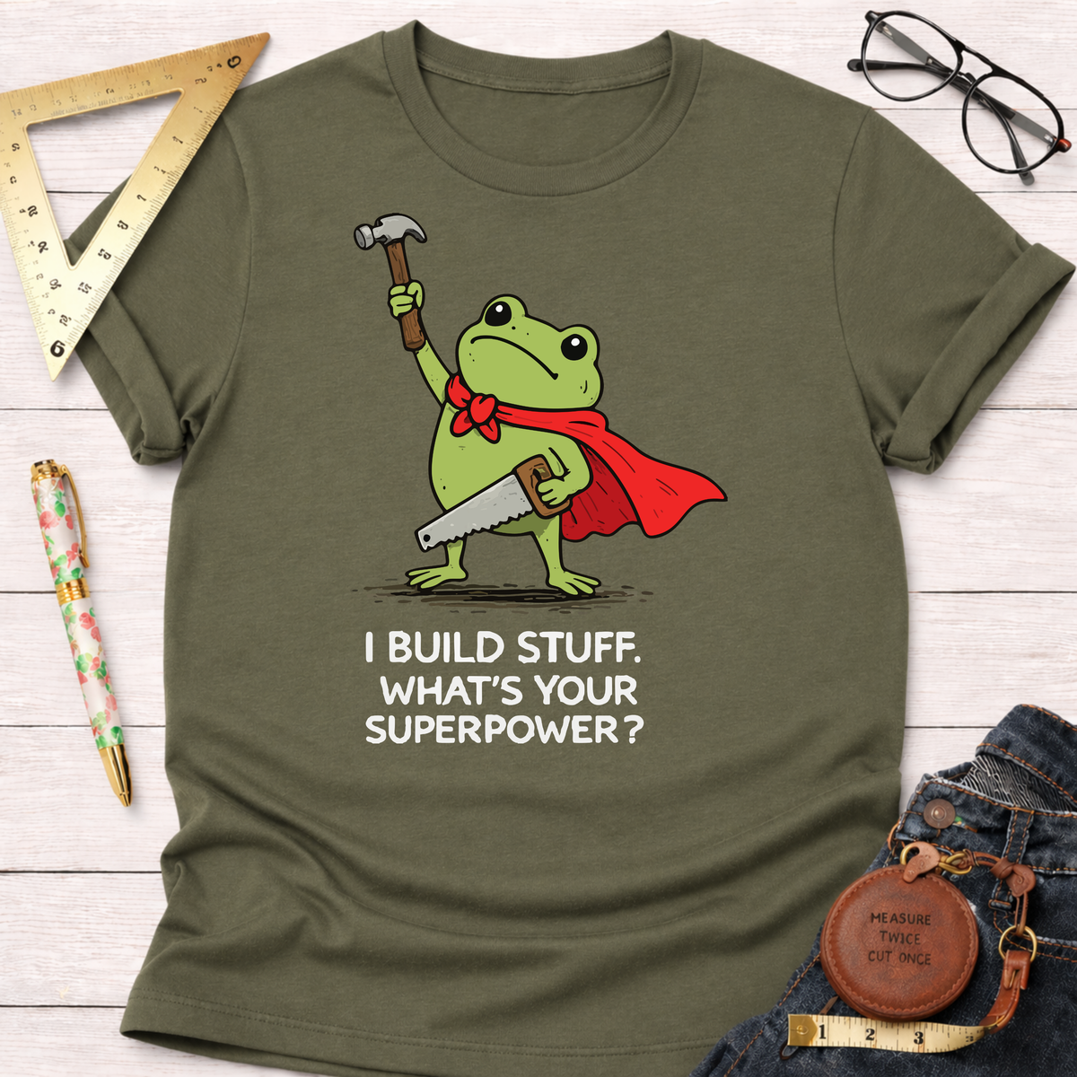 Olive green cotton tee with a frog in a red cape holding a hammer and saw, "I BUILD STUFF. WHAT'S YOUR SUPERPOWER?" text. Women's funny woodworking graphic tee shirt for female woodworkers, makers, and DIY builders — Sawdust & Ponytails