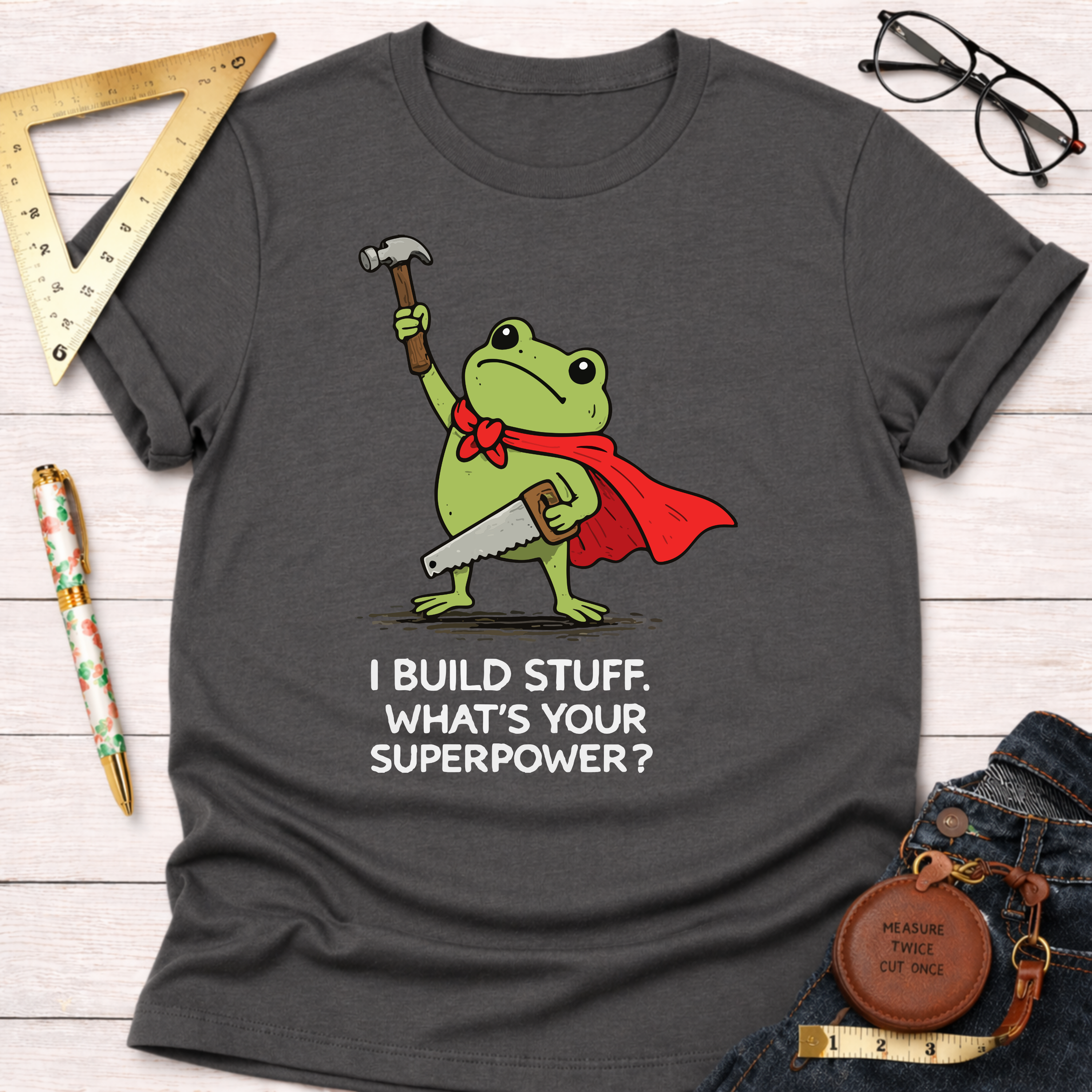 Charcoal gray cotton tee with a frog in a red cape holding a hammer and saw, "I BUILD STUFF. WHAT'S YOUR SUPERPOWER?" text. Women's funny woodworking graphic tee shirt for female woodworkers, makers, and DIY builders — Sawdust & Ponytails
