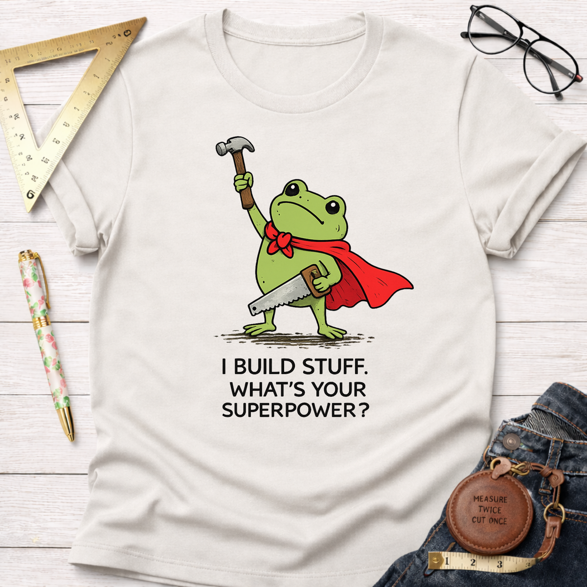 Women's funny woodworking graphic tee shirt for female woodworkers, makers, and DIY builders — Sawdust & Ponytails. Cream t-shirt with a frog superhero holding a hammer and saw, text "I BUILD STUFF. WHAT'S YOUR SUPERPOWER?".