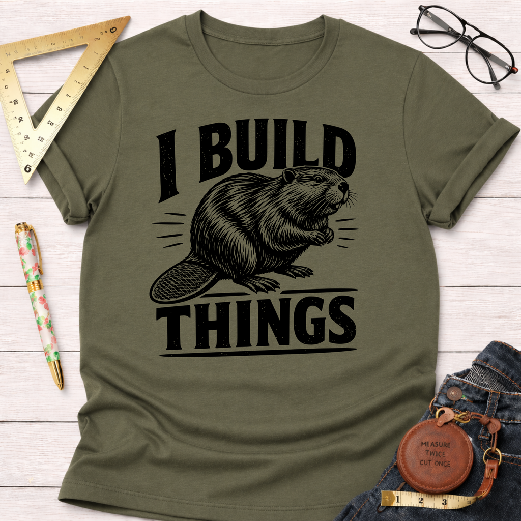Olive green "I Build Things" beaver t-shirt, folded on a white wooden surface with ruler, glasses, and jeans.