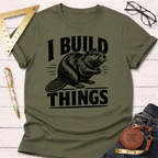 Olive green "I Build Things" beaver t-shirt, folded on a white wooden surface with ruler, glasses, and jeans.