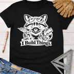 Black cotton t-shirt with a white graphic of a raccoon wearing goggles, holding a drill and saw, with "I Build Things" text. Women's funny woodworking graphic tee shirt for female woodworkers, makers, and DIY builders — Sawdust & Ponytails.