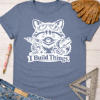 Women's funny woodworking graphic tee shirt for female woodworkers, makers, and DIY builders — Sawdust & Ponytails. Blue crewneck t-shirt with a white graphic of a raccoon wearing goggles, holding a drill and circular saw, with "I Build Things" text.