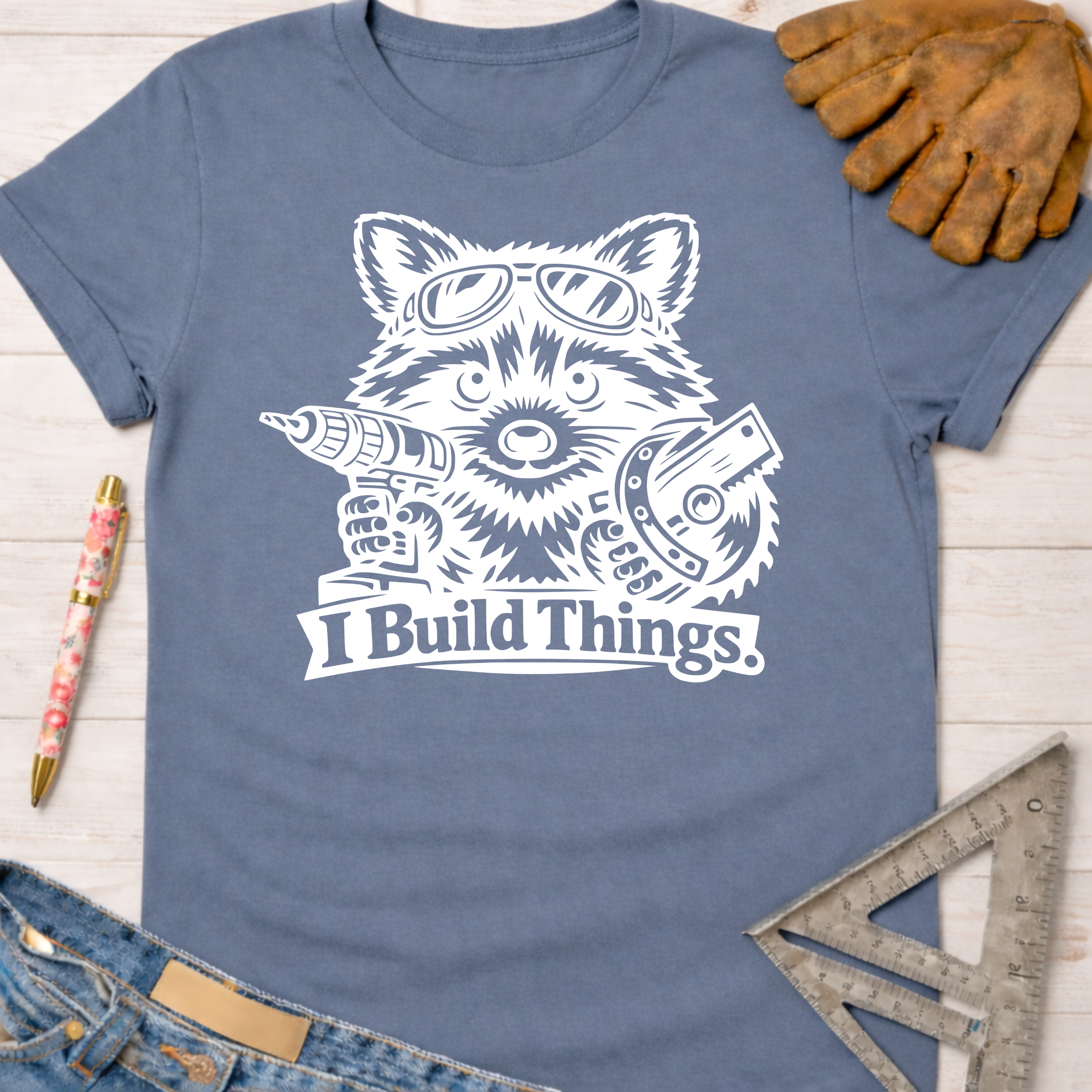 Women's funny woodworking graphic tee shirt for female woodworkers, makers, and DIY builders — Sawdust & Ponytails. Blue crewneck t-shirt with a white graphic of a raccoon wearing goggles, holding a drill and circular saw, with "I Build Things" text.