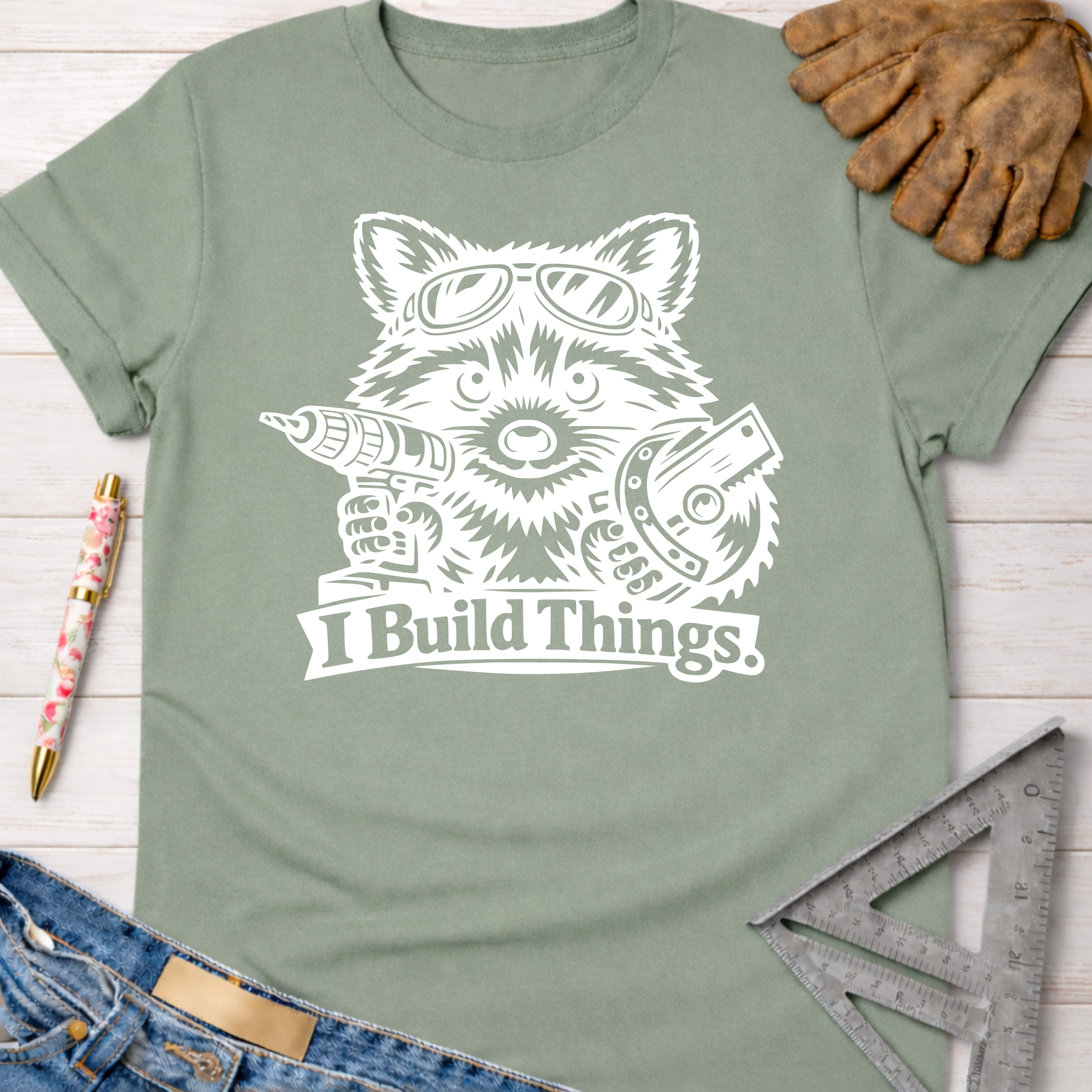 Women's funny woodworking graphic tee shirt for female woodworkers, makers, and DIY builders — Sawdust & Ponytails. Sage green cotton tee with white raccoon holding drill and saw, "I Build Things" text.
