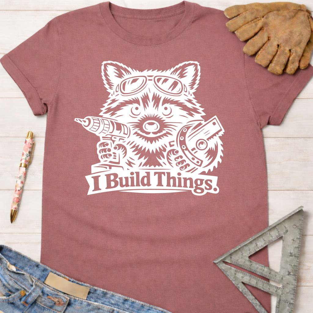 Women's funny woodworking graphic tee shirt for female woodworkers, makers, and DIY builders — Sawdust & Ponytails. Mauve cotton tee with white raccoon wearing goggles, holding a drill and saw, "I Build Things" text.