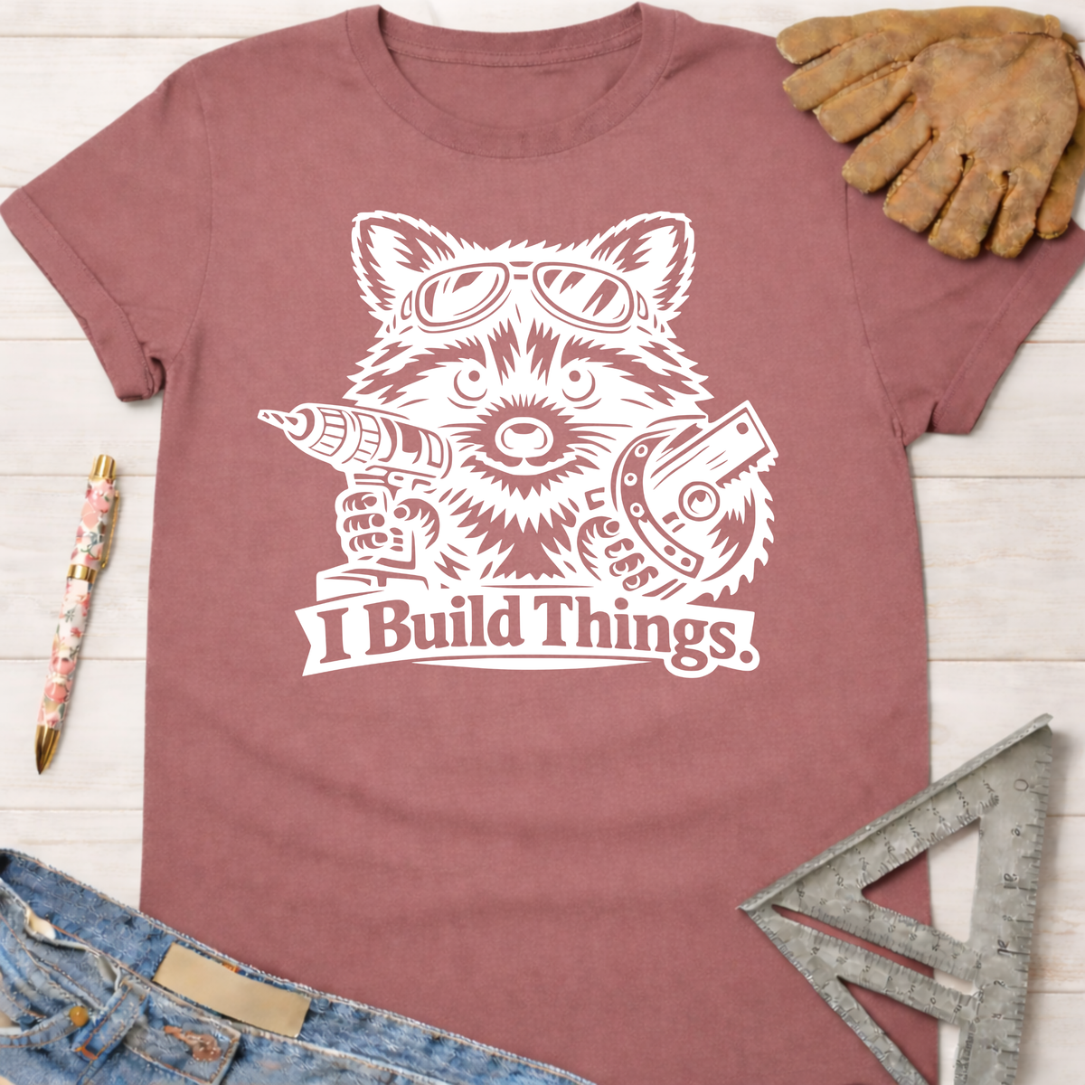 Women's funny woodworking graphic tee shirt for female woodworkers, makers, and DIY builders — Sawdust & Ponytails. Mauve cotton tee with white raccoon wearing goggles, holding a drill and saw, "I Build Things" text.