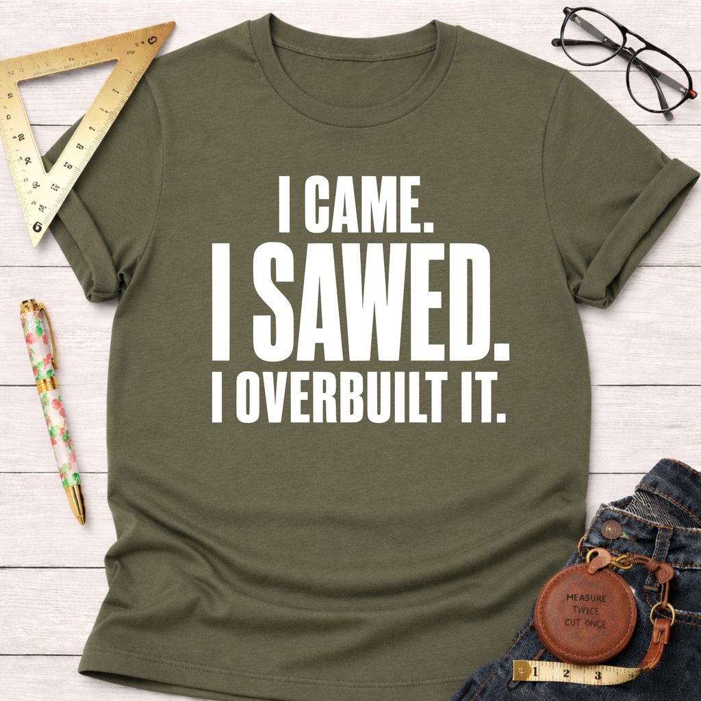 Olive green "I Came. I Sawed. I Overbuilt It." t-shirt with white text, styled on a white wooden surface.